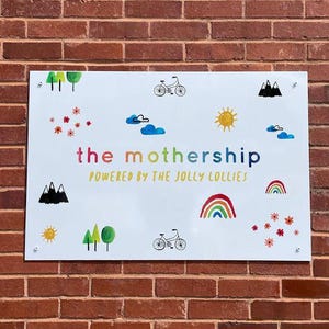 Op de afbeelding: Wit rechthoekig bord met kleurrijke illustraties en tekst. Het bord toont een regenboog, zon, wolken, bomen, bloemen en fietsen. De tekst luidt "the mothership" en "POWERED BY THE JOLLY LOLLIES". Het bord is bevestigd aan een rode bakstenen muur.