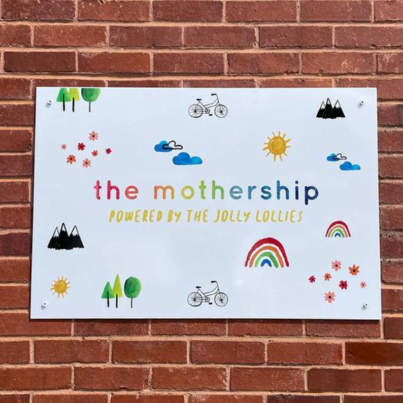 Op de afbeelding: Wit rechthoekig bord met kleurrijke illustraties en tekst. Het bord toont een regenboog, zon, wolken, bomen, bloemen en fietsen. De tekst luidt "the mothership" en "POWERED BY THE JOLLY LOLLIES". Het bord is bevestigd aan een rode bakstenen muur.