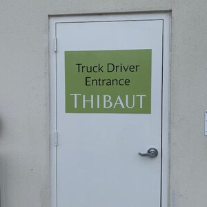 Op de afbeelding: Een witte deur met een zilveren handvat en een groen rechthoekig bord. Het bord leest "Truck Driver Entrance" boven de naam "THIBAUT" in witte hoofdletters. De deur staat in een lichtgrijze muur.