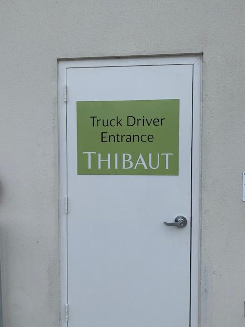 Op de afbeelding: Een witte deur met een zilveren handvat en een groen rechthoekig bord. Het bord leest "Truck Driver Entrance" boven de naam "THIBAUT" in witte hoofdletters. De deur staat in een lichtgrijze muur.