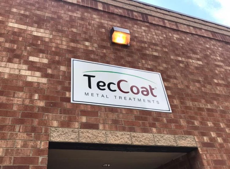 Op de afbeelding: Een bakstenen gebouw met een bord met de tekst "TecCoat METAL TREATMENTS". Het bord is wit met zwarte en bordeauxrode letters en een groene gebogen lijn. Boven het bord is een rechthoekige lamp gemonteerd.
