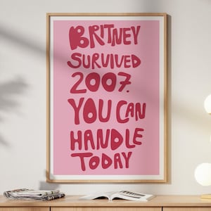 May include: A framed wall art print featuring a pink background with bold, dark red, hand-lettered text that reads: BRITNEY SURVIVED 2007. YOU CAN HANDLE TODAY. The print is displayed in a light-colored wooden frame hanging on a plain wall.