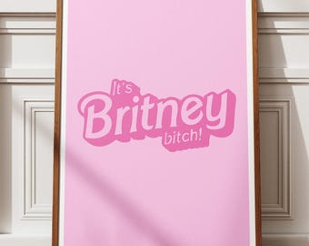 May include: A framed pink poster with the text "It's Britney bitch!" in a bubble font. The poster is in a brown wooden frame and leans against a white wall and a wooden floor.