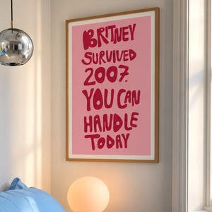 May include: A rectangular wall poster in a light wood frame featuring a pink background with bold, dark red hand-lettered text that reads: BRITNEY SURVIVED 2007. YOU CAN HANDLE TODAY. The poster is displayed on a plain white wall.