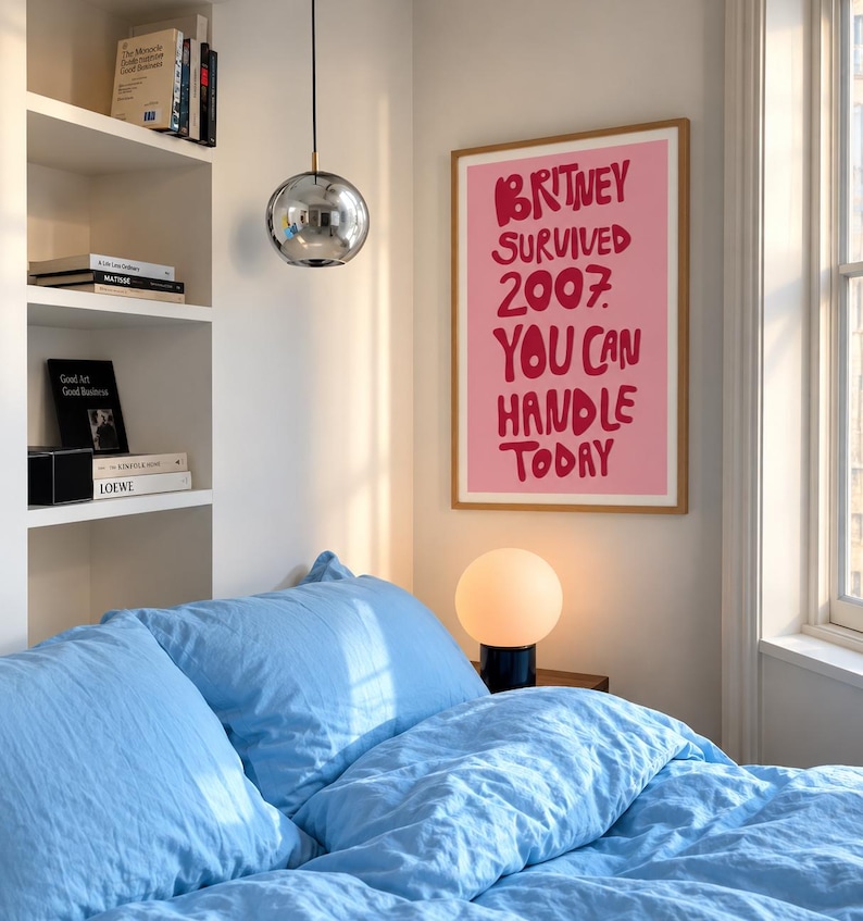 May include: A rectangular wall poster in a light wood frame featuring a pink background with bold, dark red hand-lettered text that reads: BRITNEY SURVIVED 2007. YOU CAN HANDLE TODAY. The poster is displayed on a plain white wall.