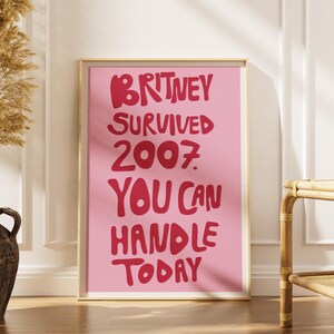 May include: A rectangular wall art print in a light gold frame featuring a pink background with bold red text that reads: BRITNEY SURVIVED 2007. YOU CAN HANDLE TODAY. The print is displayed in a vertical orientation with a clean, modern aesthetic.