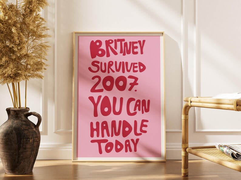 May include: A rectangular wall art print in a light gold frame featuring a pink background with bold red text that reads: BRITNEY SURVIVED 2007. YOU CAN HANDLE TODAY. The print is displayed in a vertical orientation with a clean, modern aesthetic.