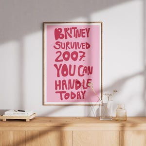 May include: A rectangular wall print in a wooden frame featuring a light pink background with bold, dark pink text that reads: BRITNEY SURVIVED 2007. YOU CAN HANDLE TODAY. The lettering is stylized in a rounded, playful font centered on the poster.