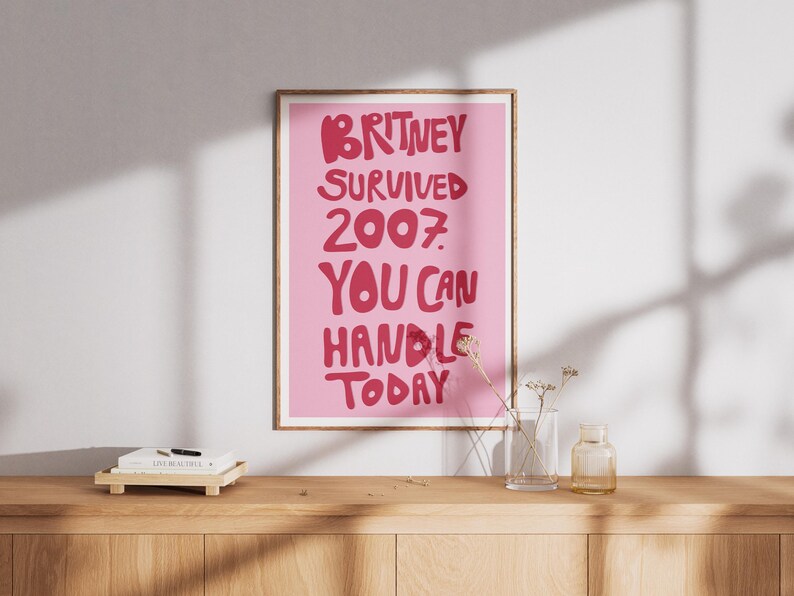 May include: A rectangular wall print in a wooden frame featuring a light pink background with bold, dark pink text that reads: BRITNEY SURVIVED 2007. YOU CAN HANDLE TODAY. The lettering is stylized in a rounded, playful font centered on the poster.