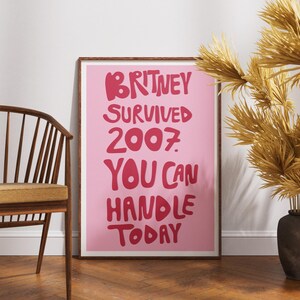 May include: A rectangular framed wall art print featuring a light pink background with bold, dark pink hand-lettered text that reads: BRITNEY SURVIVED 2007. YOU CAN HANDLE TODAY. The print is encased in a simple, thin brown wooden frame.