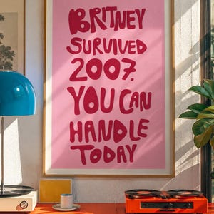 May include: A framed wall art print featuring a light pink background with bold, dark pink hand-lettered text that reads: BRITNEY SURVIVED 2007 YOU CAN HANDLE TODAY. The poster is displayed in a thin, light-colored wooden frame on a wall.