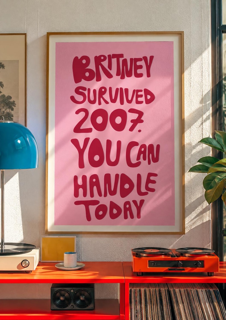 May include: A framed wall art print featuring a light pink background with bold, dark pink hand-lettered text that reads: BRITNEY SURVIVED 2007 YOU CAN HANDLE TODAY. The poster is displayed in a thin, light-colored wooden frame on a wall.