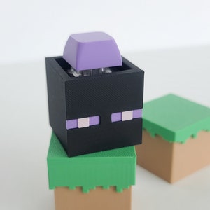 May include: A 3D printed Minecraft Creeper head with purple eyes and a purple keycap on top. The Creeper is sitting on a green and brown block.