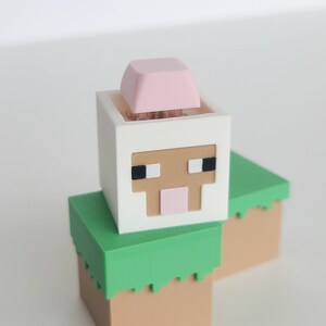 May include: A white Minecraft sheep keycap with a pink wool block on top, sitting on a green grass block and a brown dirt block.