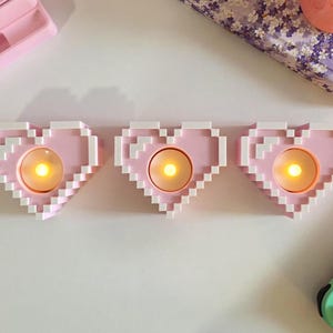 May include: Three pink pixel heart-shaped candle holders with white trim, each holding a glowing yellow candle. The candle holders are arranged in a row on a white surface. The overall theme is romantic and decorative.