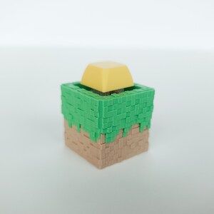 K&ouml;nnte beinhalten: Ein kleines, 3D-gedrucktes Modell eines Minecraft-Blocks mit einer gr&uuml;nen Grasdecke und einem gelben Block auf dem Gras.