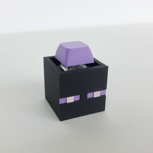 May include: A black 3D printed keycap holder shaped like the Enderman from the video game Minecraft. The holder has a purple keycap on top and purple eyes on the front.