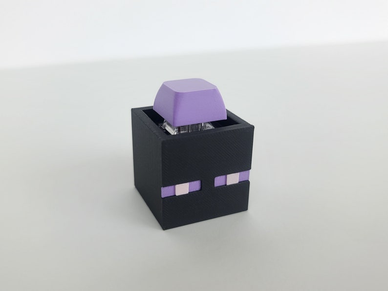 May include: A black 3D printed keycap holder shaped like the Enderman from the video game Minecraft. The holder has a purple keycap on top and purple eyes on the front.
