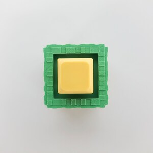 K&ouml;nnte beinhalten: Ein gelber quadratischer Knopf in einem gr&uuml;nen quadratischen Rahmen. Der Rahmen hat eine strukturierte Oberfl&auml;che.