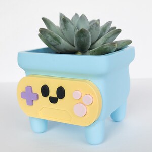 May include: A light blue planter shaped like a video game controller with a yellow face and pink buttons. The planter has four short legs and is holding a green succulent plant.