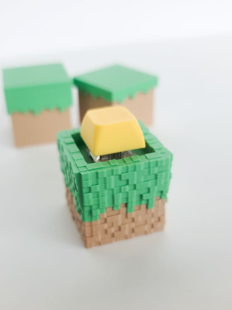 K&ouml;nnte beinhalten: Ein gelber Keycap sitzt auf einem gr&uuml;nen und braunen Block, der einem Minecraft-Block &auml;hnelt. Zwei weitere &auml;hnliche Bl&ouml;cke sind im Hintergrund sichtbar.