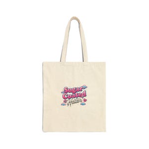 Può includere: Una borsa tote color crema con una grafica rosa, blu e bianca che recita "Sugar Coated Hater". La borsa ha due manici lunghi ed è realizzata in un materiale simile alla tela. La grafica include nuvole e un bacio.