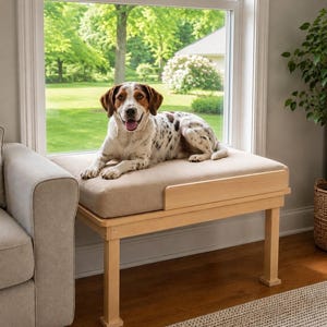 May include: A light-coloured wooden pet bench featuring a rectangular frame with four straight legs and a thick, beige upholstered cushion top. The bench is positioned directly in front of a large window, providing a raised platform for a dog.