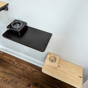 May include: Two wall-mounted pet food and water bowls. The top bowl is on a dark brown shelf, while the bottom bowl is on a light wood shelf.