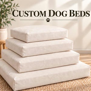 May include: A stack of four rectangular, cream-colored dog beds in varying sizes, arranged from smallest at the top to largest at the bottom. The text CUSTOM DOG BEDS is displayed above the stack, accompanied by a small paw print icon.