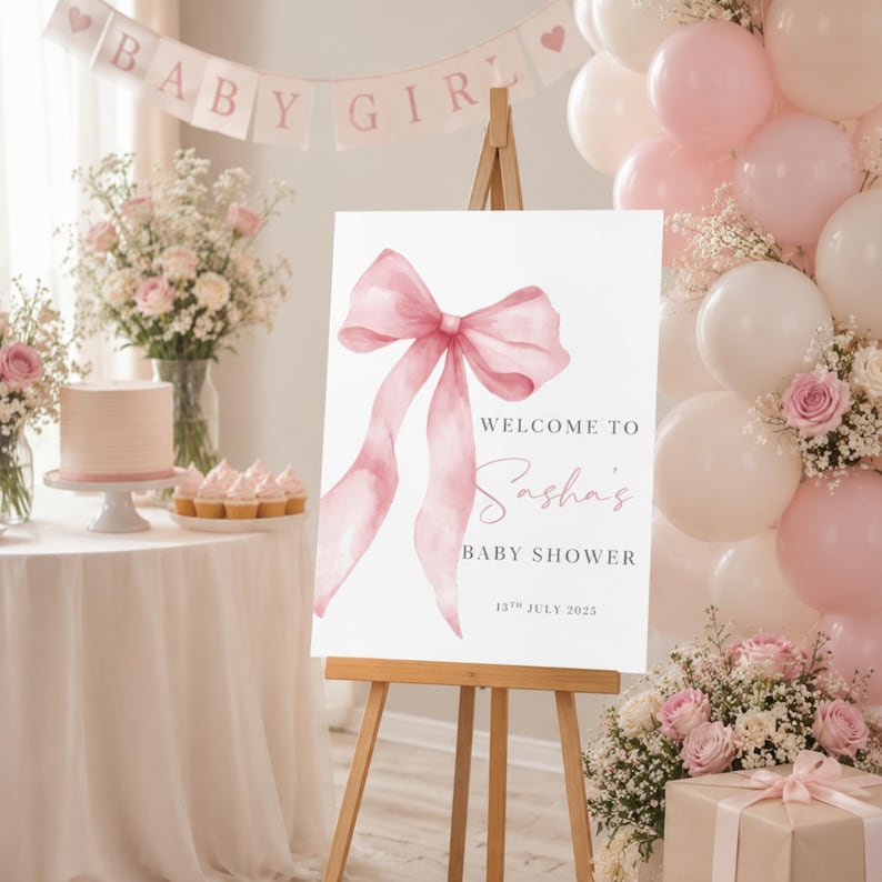 May include: A white rectangular welcome sign on a wooden easel featuring a large watercolor pink bow illustration. The text reads: Welcome to Sasha's Baby Shower, 13th July 2023. The design uses black serif and pink script fonts on a clean white background.