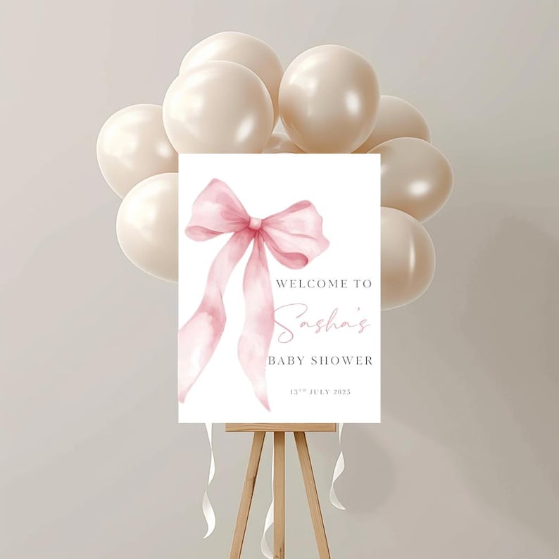 May include: A rectangular sign on a wooden easel featuring a watercolor illustration of a pink ribbon bow. The text reads: WELCOME TO Sasha's BABY SHOWER 13TH JULY 2025. The sign is surrounded by a cluster of beige balloons tied with white ribbons.
