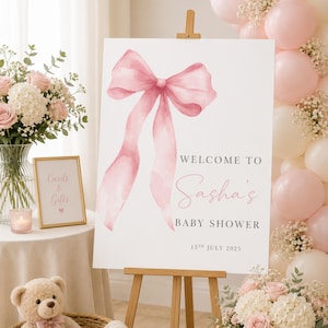 May include: A white rectangular welcome sign for a baby shower on a wooden easel, featuring a large watercolor pink bow illustration and the text: Welcome to Sasha's Baby Shower, 13th July 2025. The sign is displayed with pink balloons and flowers.