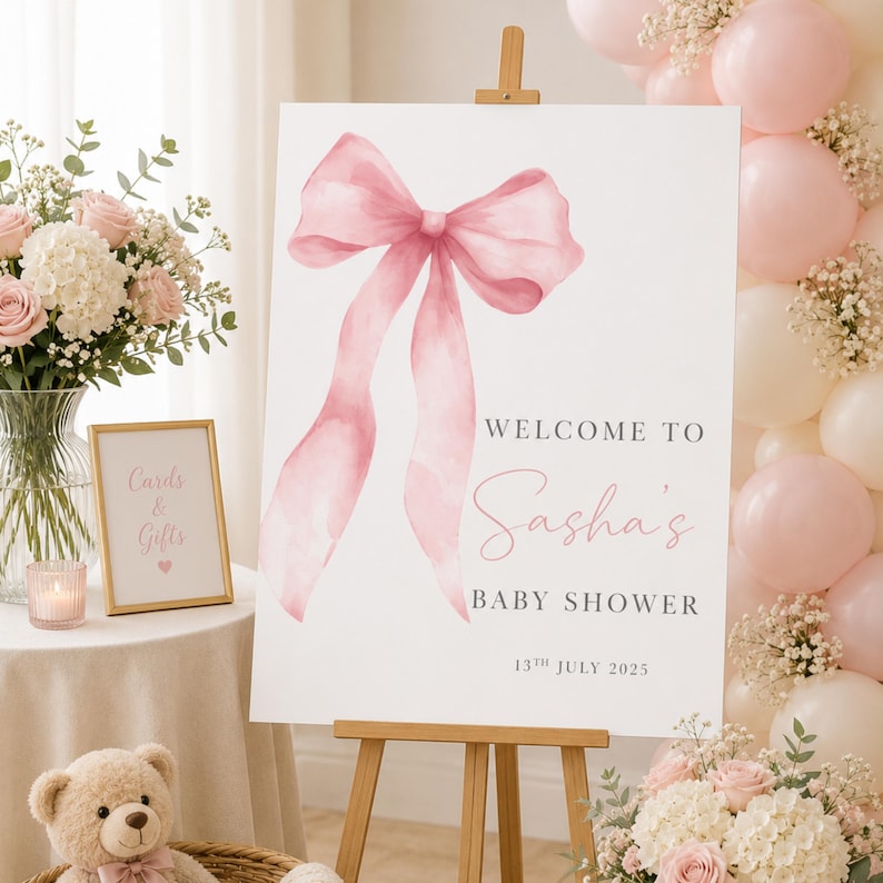 May include: A white rectangular welcome sign for a baby shower on a wooden easel, featuring a large watercolor pink bow illustration and the text: Welcome to Sasha's Baby Shower, 13th July 2025. The sign is displayed with pink balloons and flowers.