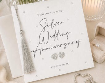 May include: A square white greeting card featuring the text "WITH LOVE ON YOUR Silver Wedding Anniversary" and "LIZ AND MARK". The card is decorated with small clear crystals, a silver tassel, and two heart-shaped stud earrings.