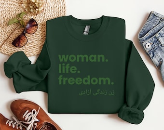 Puede incluir: Una sudadera verde oscuro con las palabras "woman. life. freedom." impresas en una fuente verde más clara. Debajo del texto en inglés hay texto en otro idioma. La sudadera se muestra sobre una superficie blanca con otros artículos.