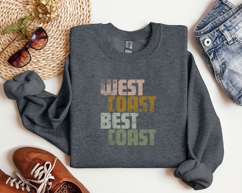 Może przedstawiać: Szara bluza z nadrukiem "West Coast Best Coast" w stylu vintage. Tekst jest w odcieniach r&oacute;żowego, ż&oacute;łtego, pomarańczowego i zielonego.
