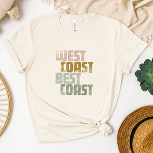 Może przedstawiać: Biała koszulka z nadrukiem "West Coast Best Coast" w stylu vintage w kolorze r&oacute;żowym, zielonym i złotym.
