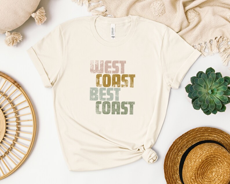 Może przedstawiać: Biała koszulka z nadrukiem "West Coast Best Coast" w stylu vintage w kolorze r&oacute;żowym, zielonym i złotym.