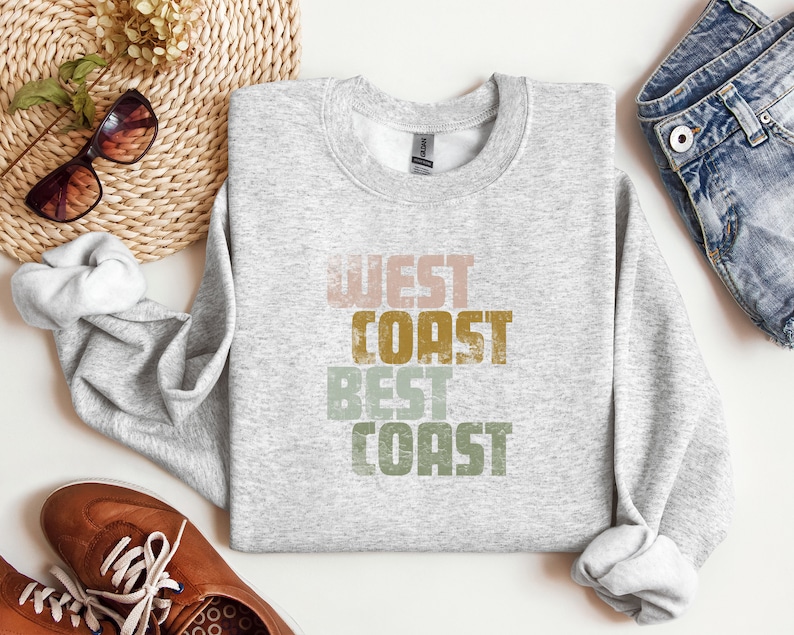 Może przedstawiać: Szara bluza z nadrukiem "West Coast Best Coast" w stylu vintage. Tekst jest w odcieniach r&oacute;żowego, ż&oacute;łtego, zielonego i brązowego.