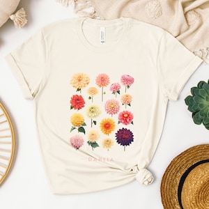 Puede incluir: Una camiseta de color crema con un estampado gráfico de una variedad de flores de dalia de colores. El texto "DAHLIA" está impreso debajo de las flores.
