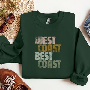 Może przedstawiać: Zielony sweter z nadrukiem "West Coast Best Coast" w stylu vintage. Tekst jest w odcieniach brązu, ż&oacute;łtego i białego.