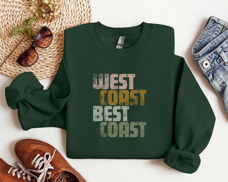 Może przedstawiać: Zielony sweter z nadrukiem "West Coast Best Coast" w stylu vintage. Tekst jest w odcieniach brązu, ż&oacute;łtego i białego.