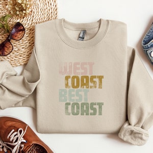 Może przedstawiać: Beżowa bluza z nadrukiem "West Coast Best Coast" w stylu vintage. Tekst jest w odcieniach r&oacute;żowego, zielonego, ż&oacute;łtego i brązowego.