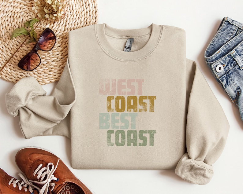 Może przedstawiać: Beżowa bluza z nadrukiem "West Coast Best Coast" w stylu vintage. Tekst jest w odcieniach r&oacute;żowego, zielonego, ż&oacute;łtego i brązowego.
