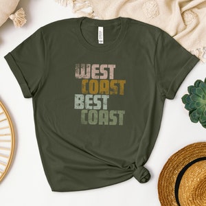 Może przedstawiać: Zielony T-shirt z napisem "West Coast Best Coast" w stylu vintage. Tekst jest w odcieniach r&oacute;żowego, ż&oacute;łtego i zielonego.