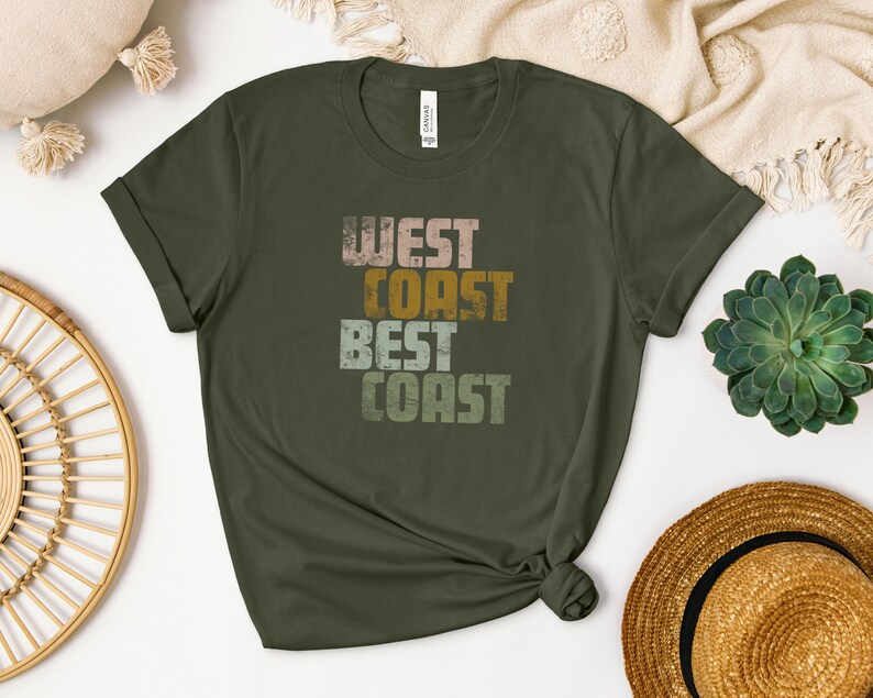 Może przedstawiać: Zielony T-shirt z napisem "West Coast Best Coast" w stylu vintage. Tekst jest w odcieniach r&oacute;żowego, ż&oacute;łtego i zielonego.