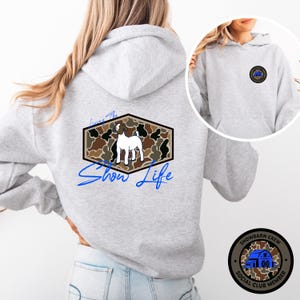May include: A light grey hoodie with a front pocket and drawstring hood. The back features a hexagon design with a goat illustration and the words "Living The Show Life." A small circular patch on the front reads "Showbarn Crew Social Club Member."