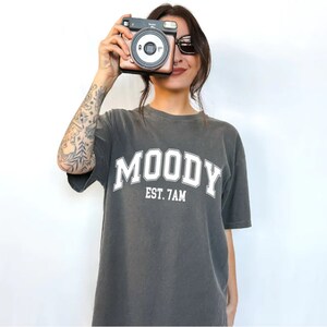 May include: A dark gray t-shirt with the text "MOODY EST. 7AM" printed in white. The shirt has a relaxed fit and a crew neck.