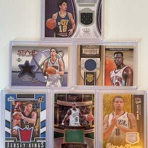 May include: A collection of six basketball trading cards in protective cases. The cards feature images of basketball players, team logos, and jersey swatches. The cards are from various brands, including Upper Deck, Donruss, and Fleer.
