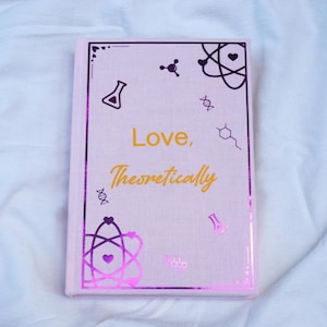 Puede incluir: Un libro rosa claro con el título "Love, Theoretically" en letras doradas. La portada presenta ilustraciones moradas con temática científica y un borde decorativo. El libro está cerrado y sobre una superficie blanca.
